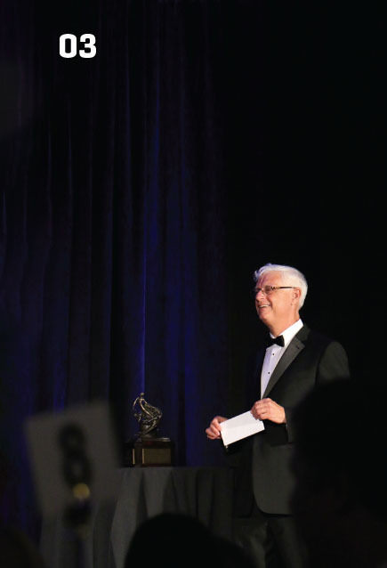 Greg Principato smiles while presenting at the 2021 Robert J. Collier Trophy Dinner. He is standing next to a small copy of the trophy 